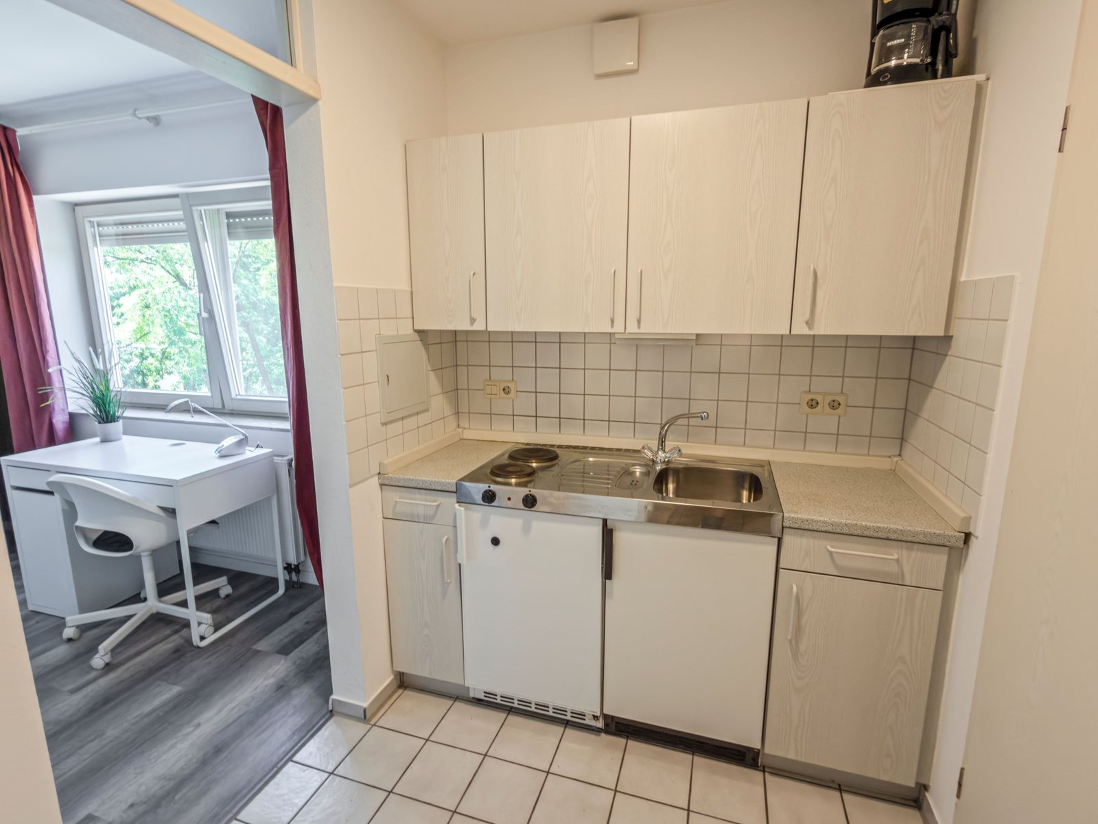 Küchenzeile mit Spüle, zwei Kochplatten, Kühlschrank und Kaffeemaschine, daneben der Wohnbereich mit Schreibtisch