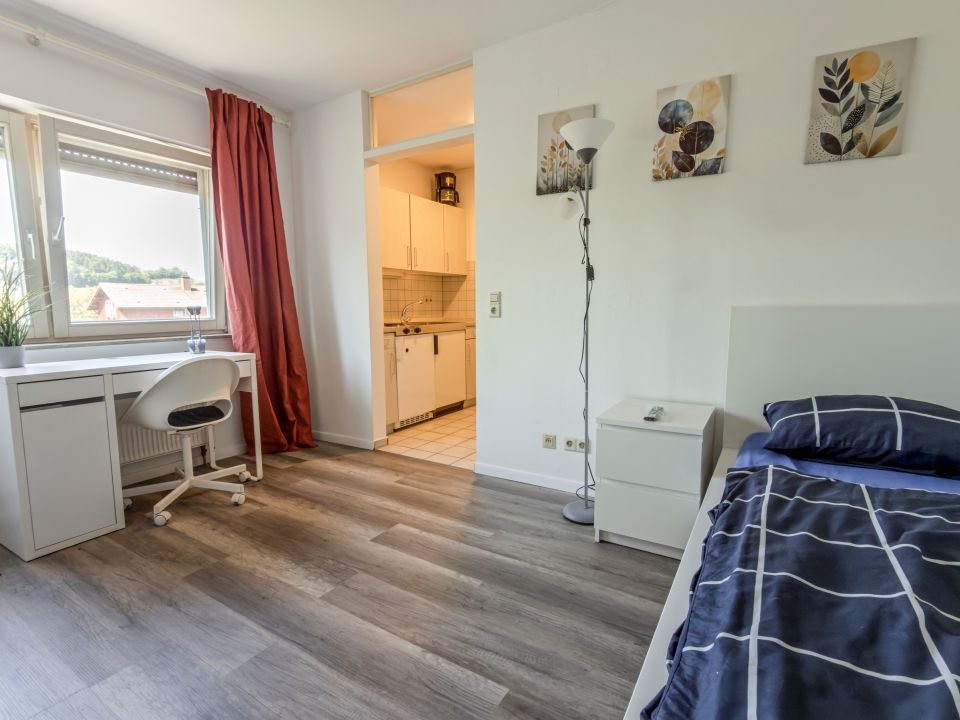 Quartier mit Bett, Schreibtisch und Bürostuhl am Fenster, dahinter die Küchenzeile